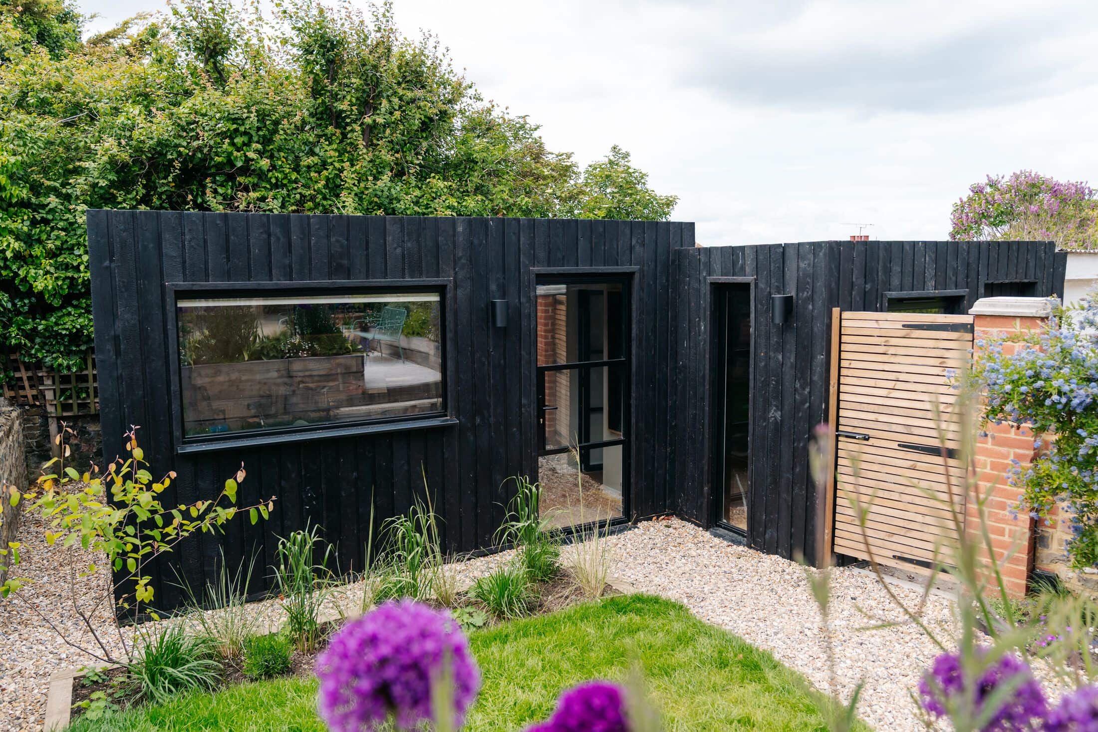Black charred-timber garden studio with full-height Crittall-style glazing, set on a gravel path among grasses and purple alliums