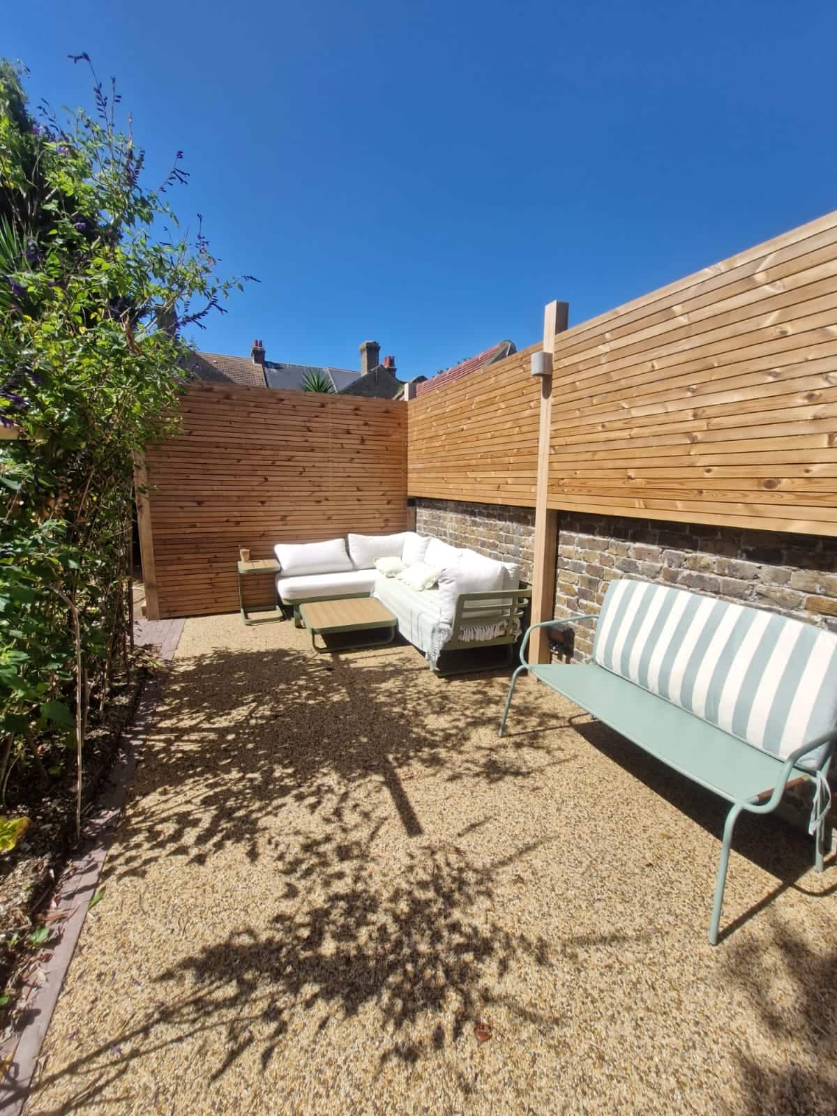 Sunny resin-bound gravel courtyard enclosed by slatted timber screens, with a striped garden bench and outdoor sofa