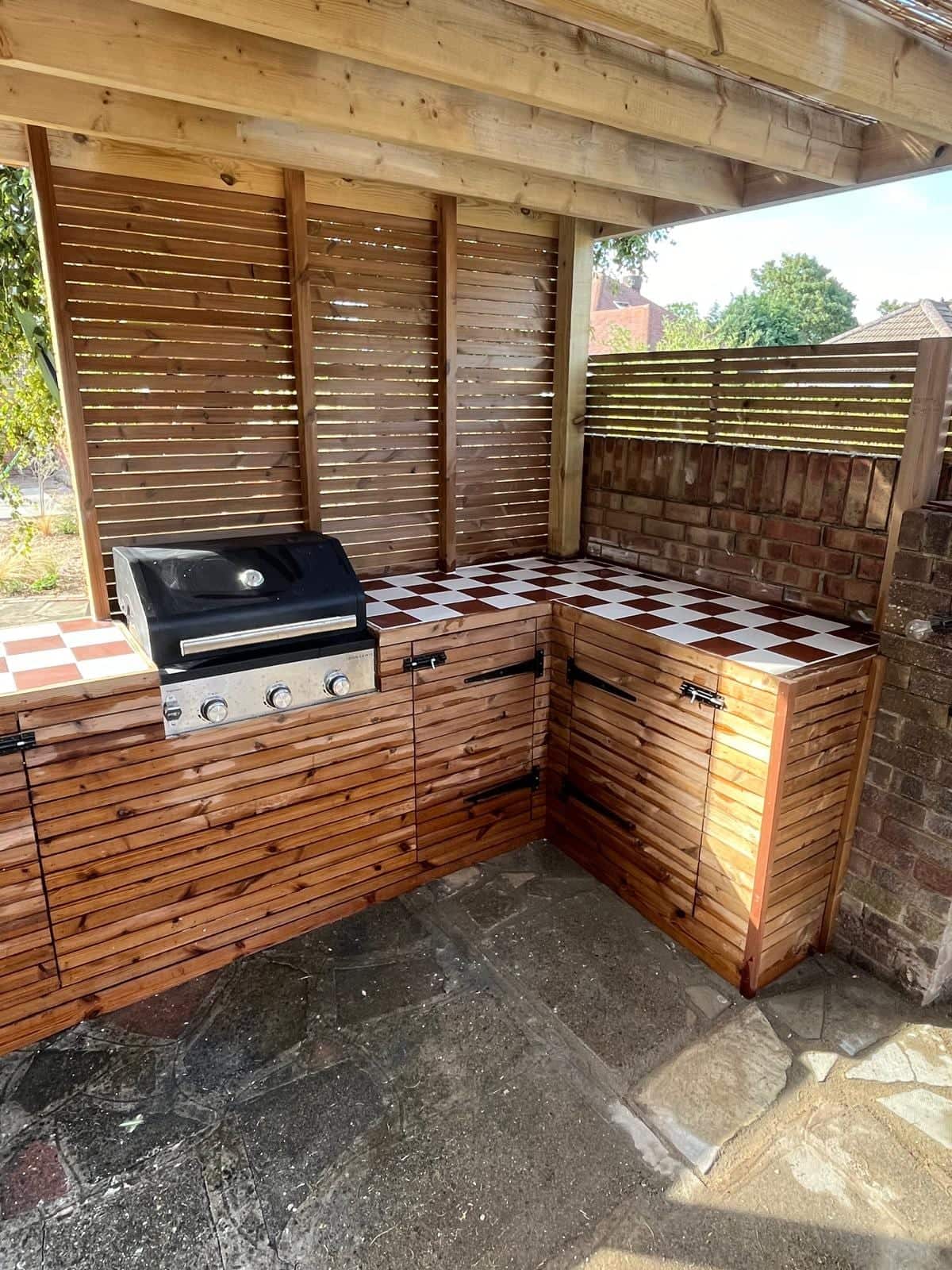 L-shaped outdoor kitchen with a built-in gas barbecue, chequered tiled worktop and slatted timber doors under a pergola