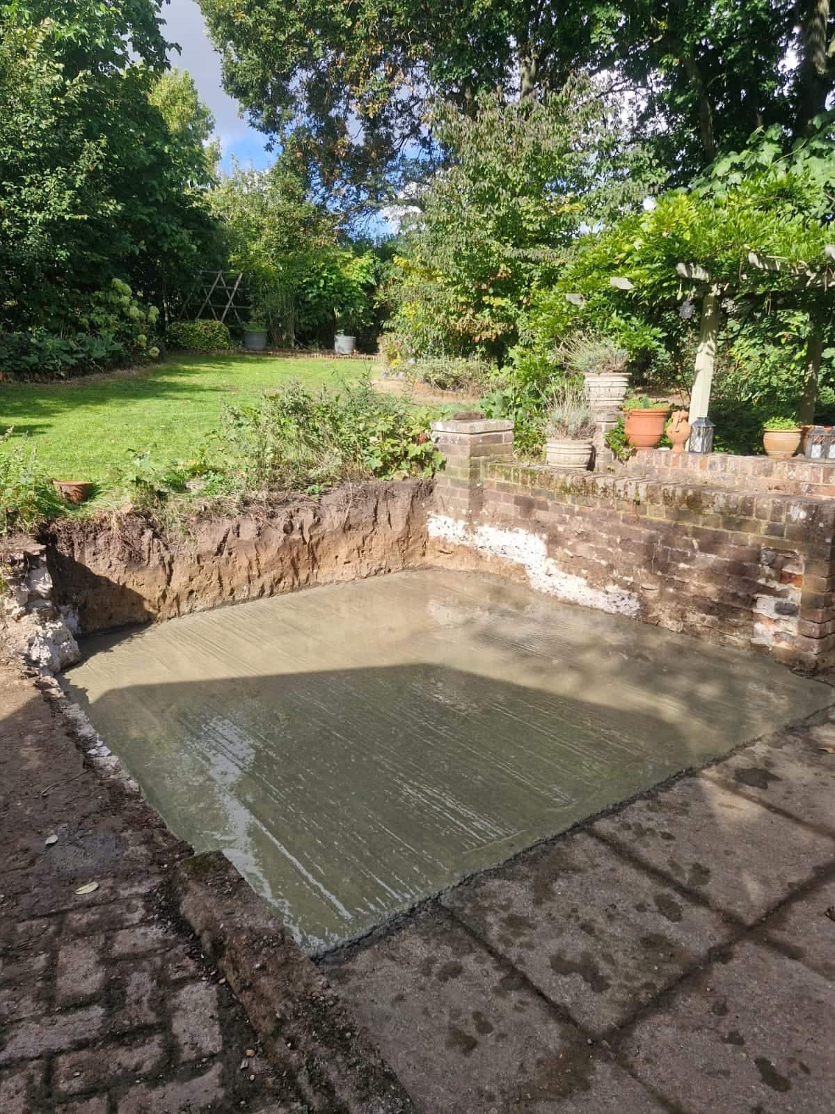 Freshly poured concrete base set into a sunken area of a mature garden, ready for a garden building