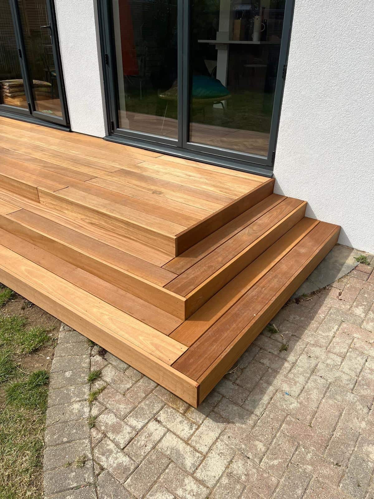 Corner detail of a new hardwood deck with stepped edges beside grey bi-fold doors and a block-paved path