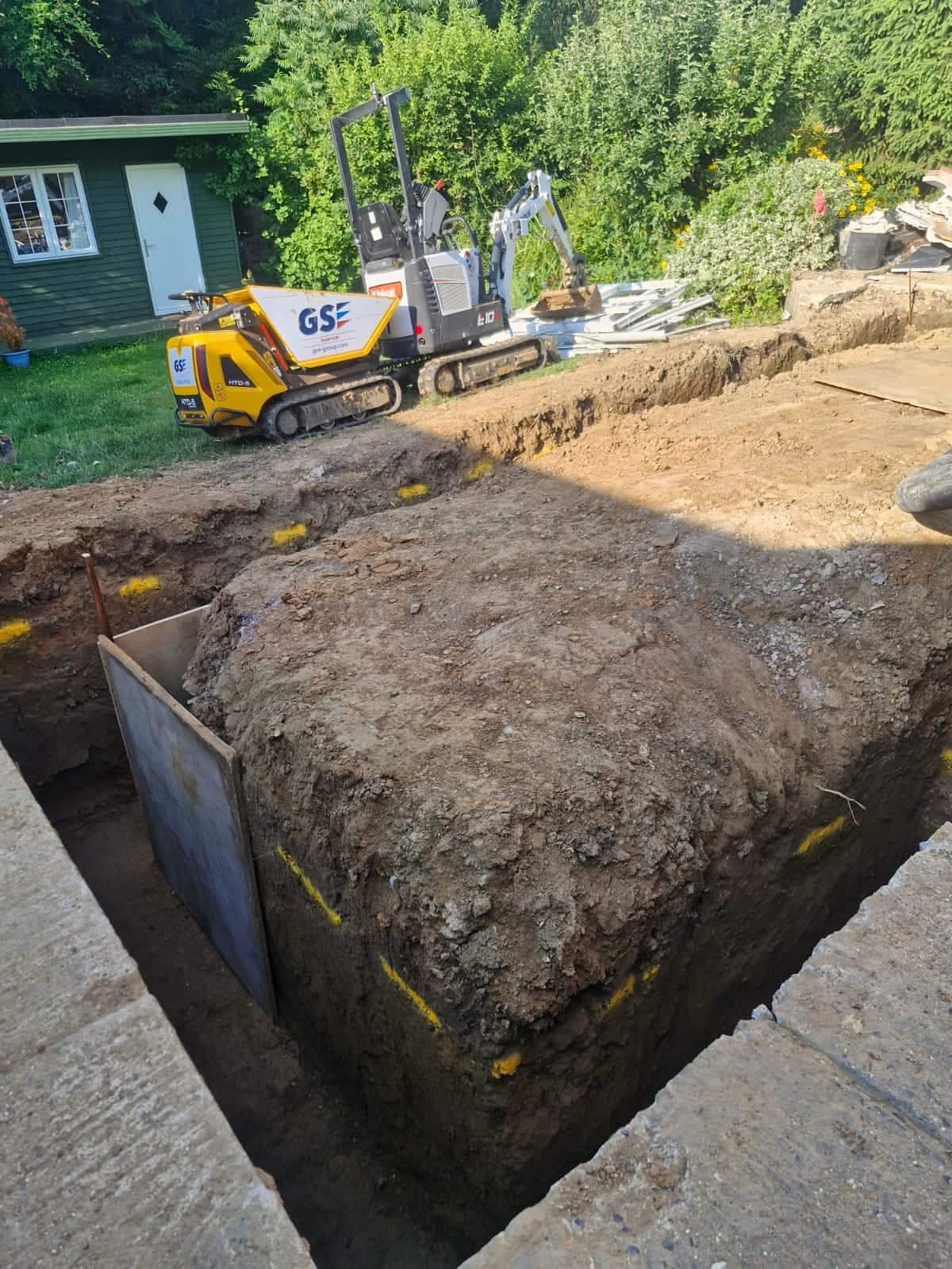Foundation trench dug and marked out for concrete, with a mini digger and tracked dumper on the lawn behind
