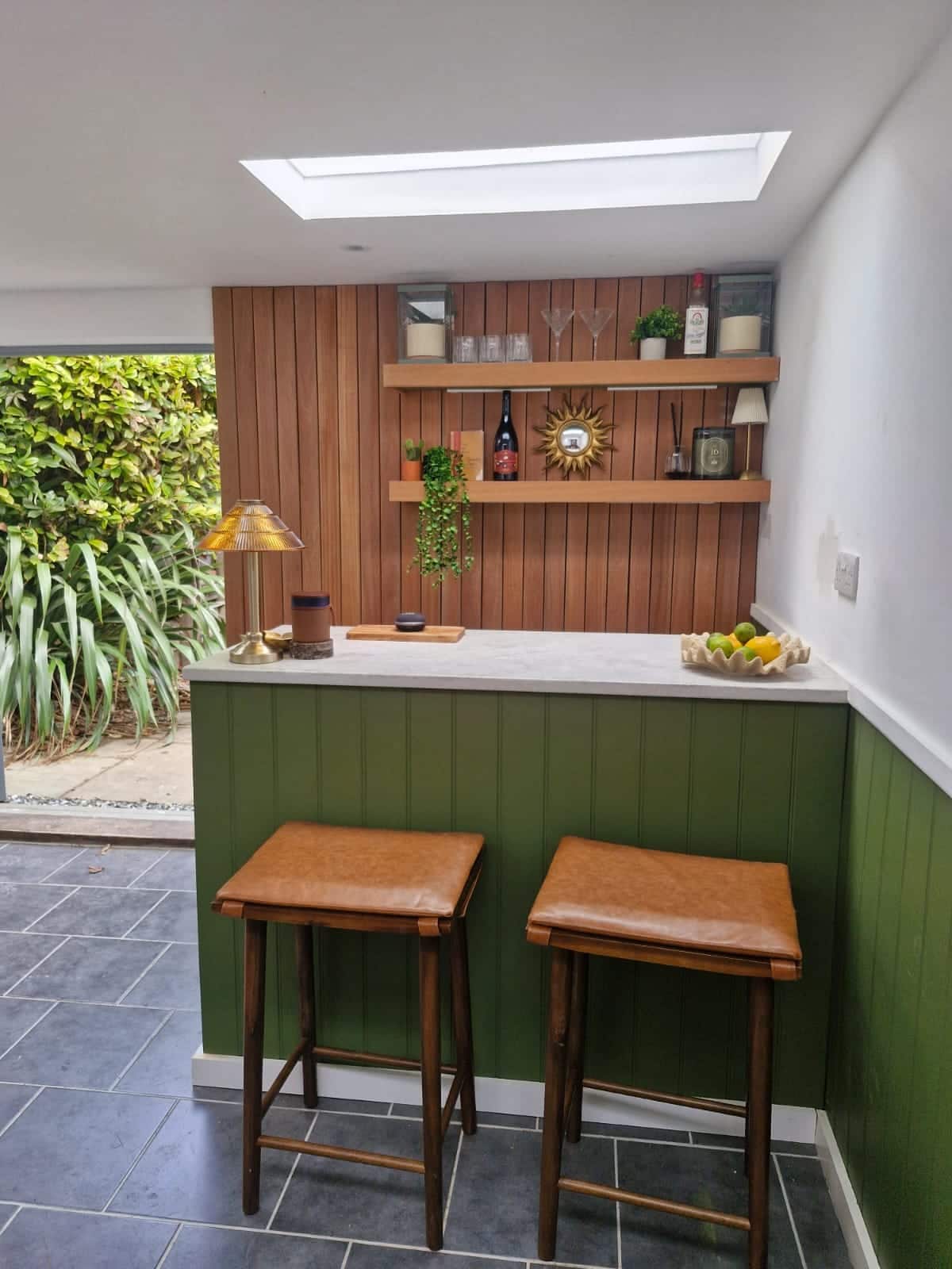 Bespoke garden bar with green panelling, floating timber shelves and a stone worktop, opening onto the garden