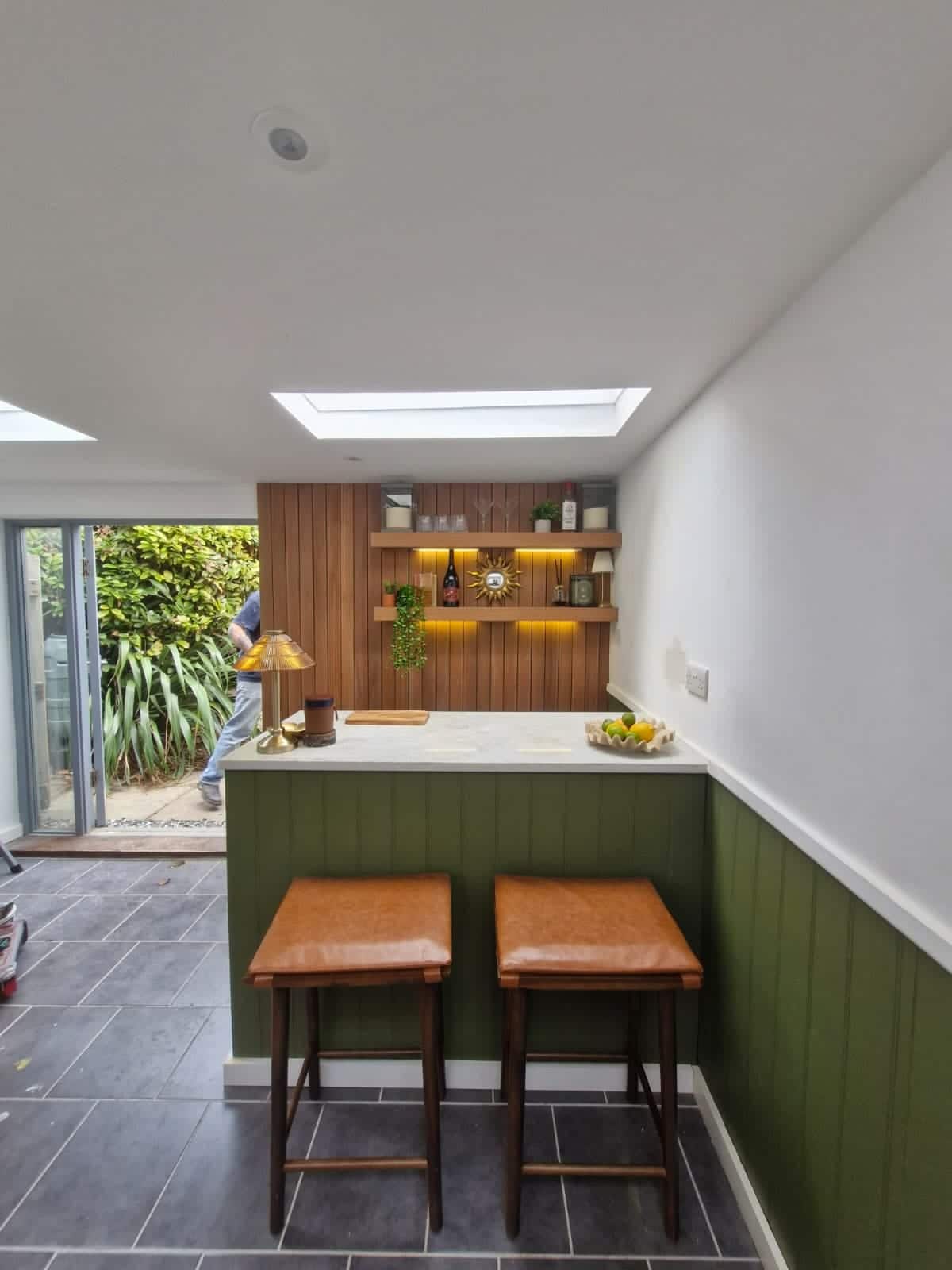 Garden room bar under a skylight with green panelled front, slatted timber feature wall and bi-fold doors to the garden