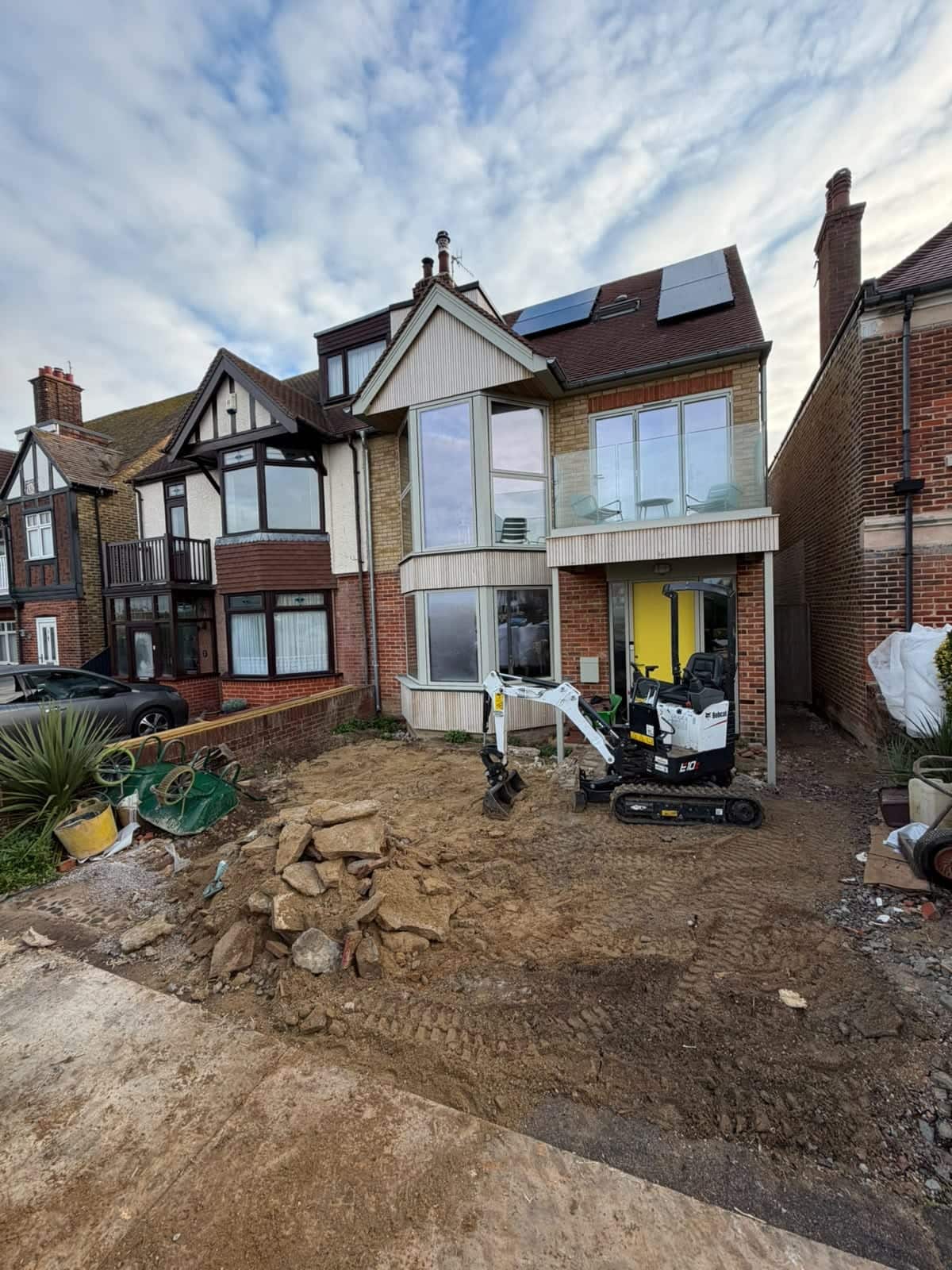 Mini digger excavating a front garden in front of a house with a new timber-clad extension and balcony