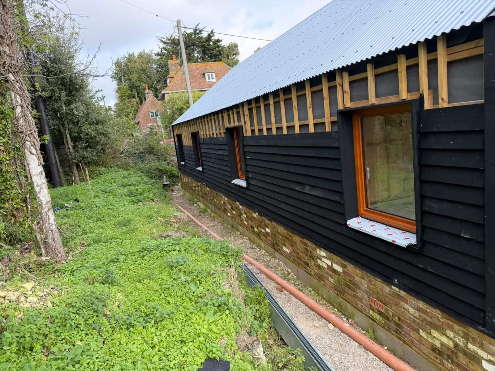 Black timber cladding being fitted to a long outbuilding with timber-framed windows and a metal roof, during construction