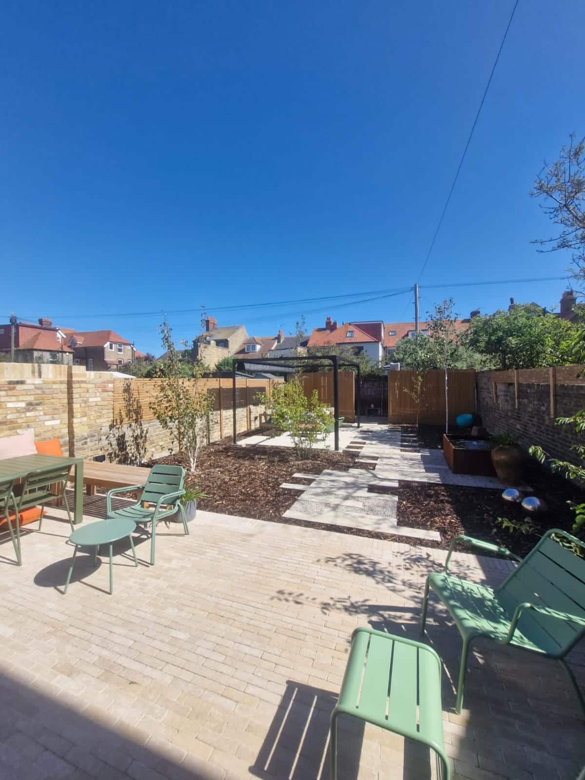 Landscaped town garden with limestone sett paving, a black steel pergola, bark-mulched beds, paving-slab stepping stones and a corten water feature