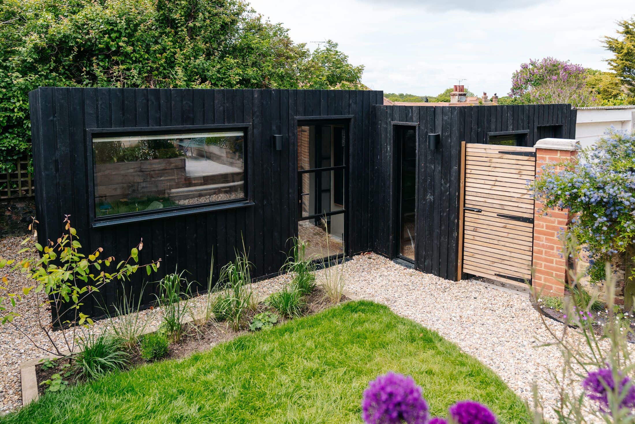 Black charred-timber garden studio with Crittall-style glazing and a slatted cedar side gate, set behind a lawn and planted borders