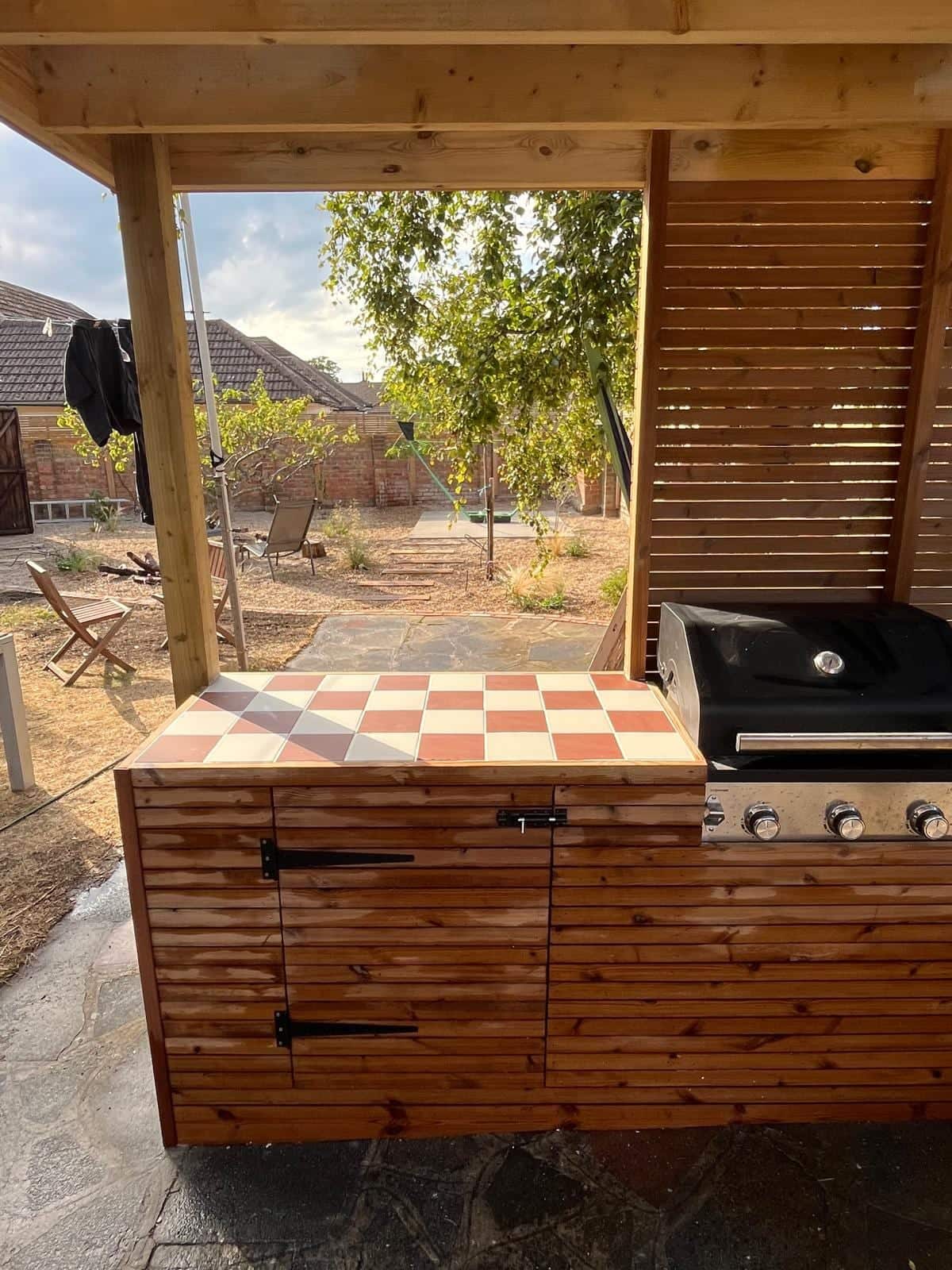 Close view of an outdoor kitchen worktop in red and white tiles with a built-in barbecue and slatted timber cupboards